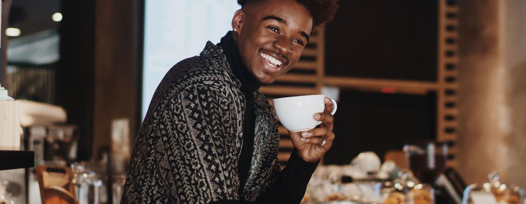 young man smiles while holding a cup of coffee in coffee shop